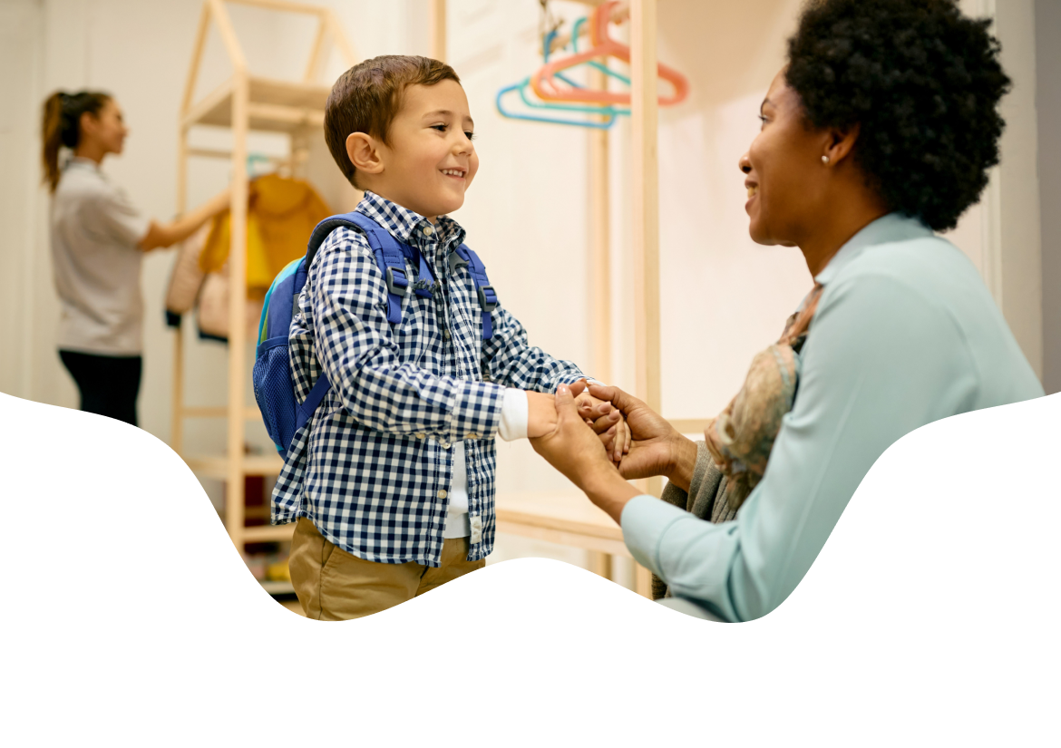 A childcare teacher greeting a smiling boy wearing a backpack