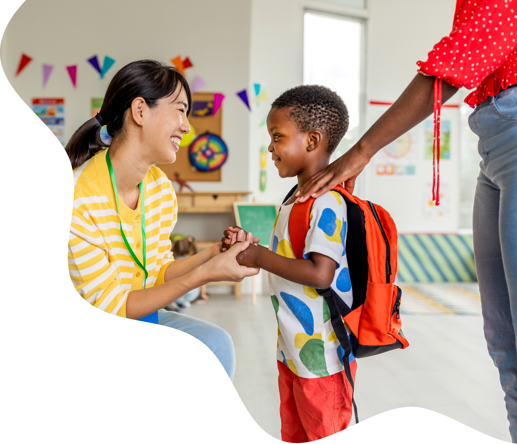 A childcare teacher greeting a child with a backpack as a parent looks on