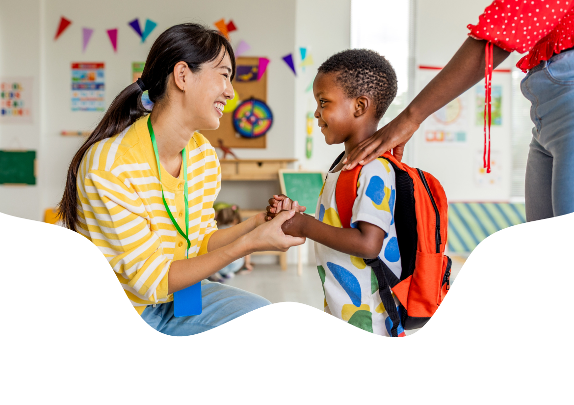 A childcare teacher greeting a child with a backpack as a parent looks on
