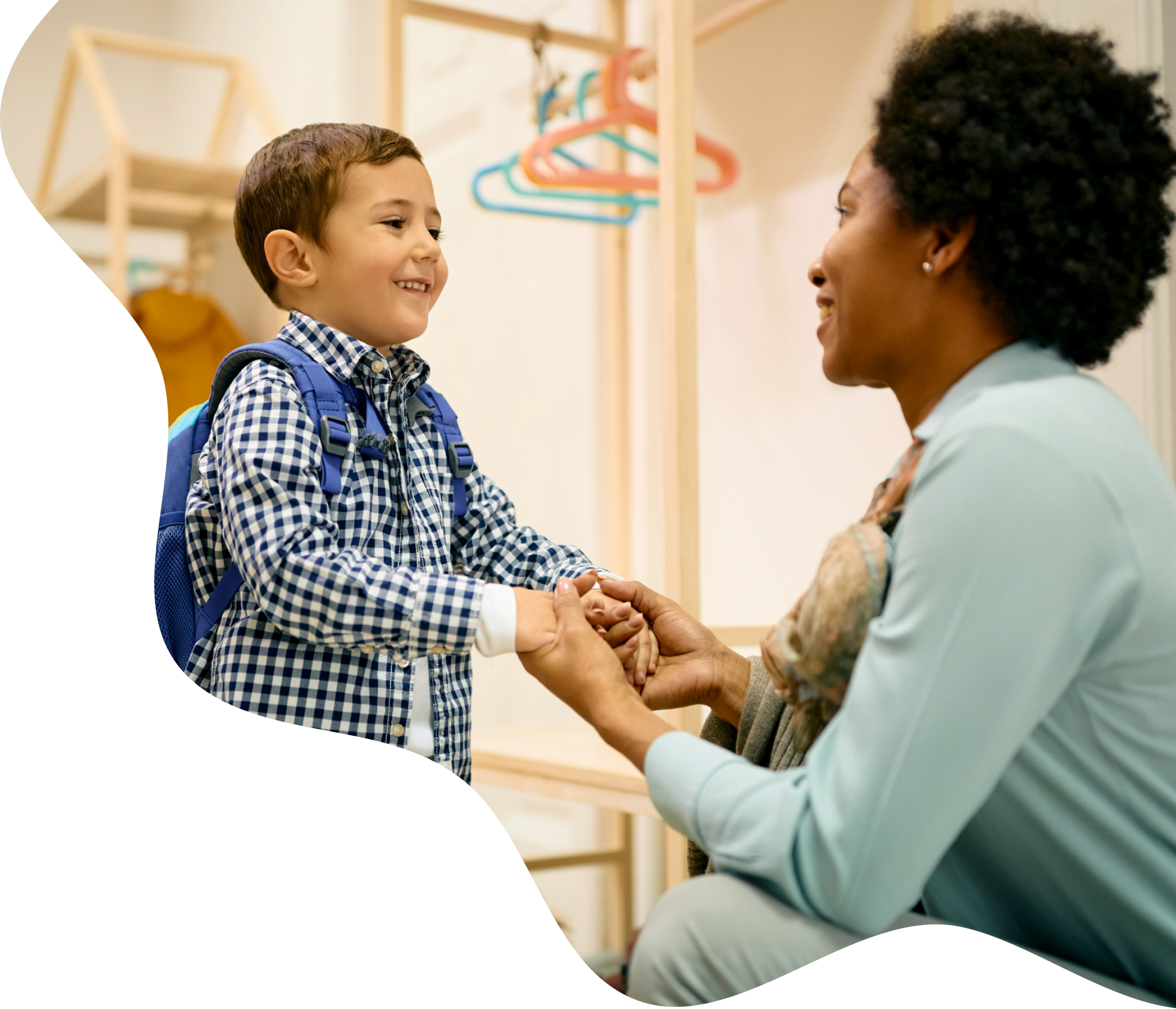 A childcare teacher kneeling to greet a smiling boy wearing a backpack